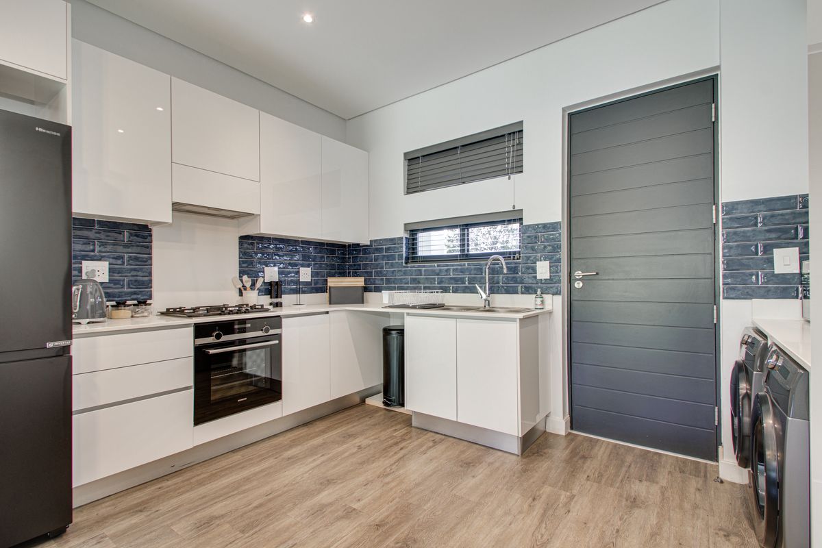 Modern kitchen featuring white high-gloss cabinetry, blue subway tile backsplash, gas hob, electric oven, and integrated laundry space with wood-look laminate flooring.