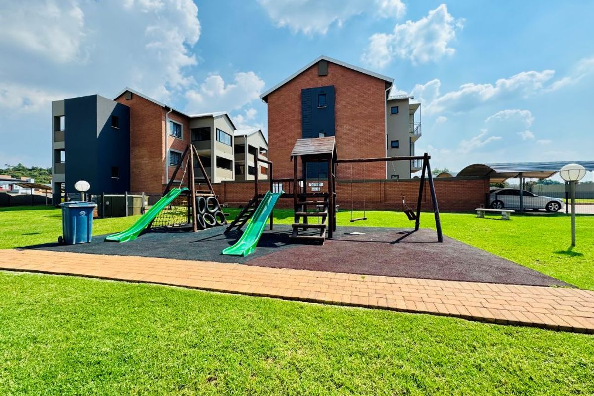 A well-maintained communal garden area featuring a children's playground, paved walkways, and adjacent apartment building blocks with covered parking.