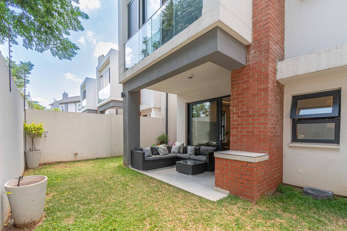A private rear garden featuring a covered patio with tiled flooring, brick feature pillar, and glass balcony railing above.
