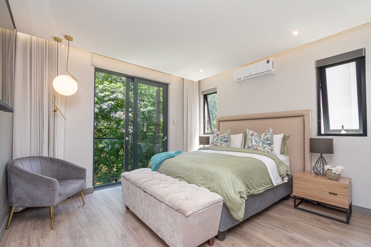 A modern, well-lit bedroom featuring wood-look flooring, a wall-mounted air conditioning unit, and large sliding glass doors providing natural light and garden views.