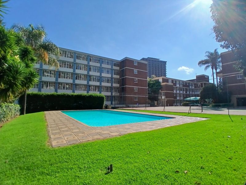 A well-maintained communal swimming pool area set within a landscaped garden, featuring paved surrounds and views of the apartment complex buildings.