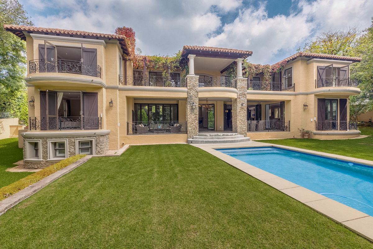 A grand Mediterranean-style rear elevation featuring a swimming pool, manicured lawn, expansive balconies with wrought-iron railings, and stone-clad architectural columns.