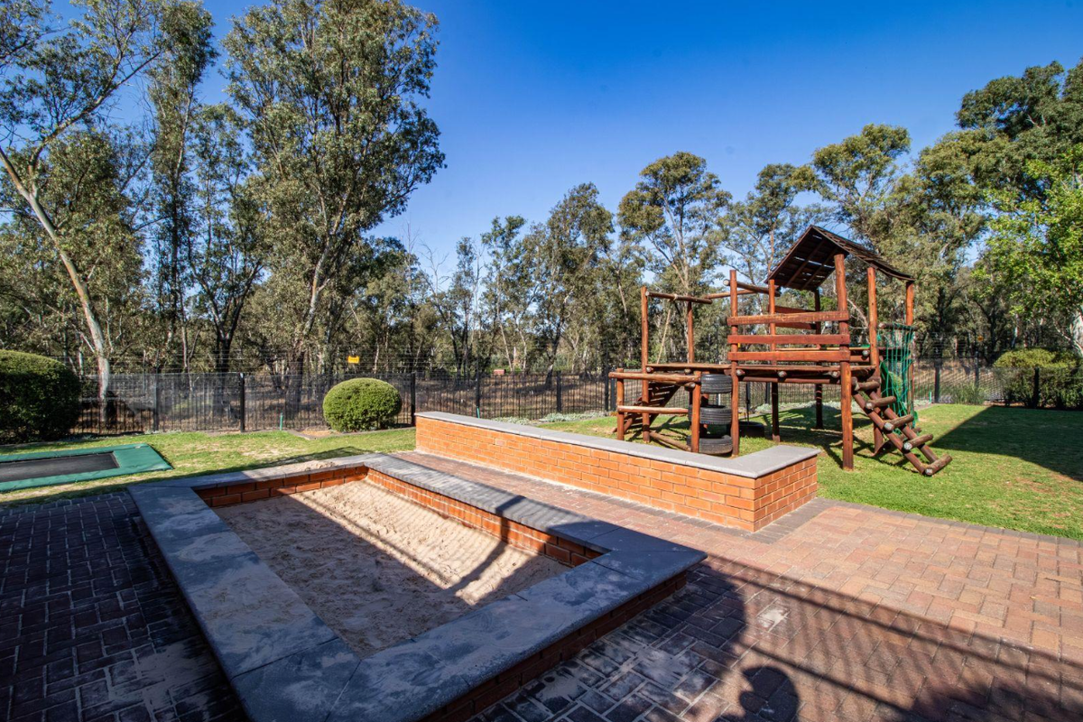 A spacious rear garden featuring a paved patio area, a built-in brick sandpit, and a wooden jungle gym set on a well-maintained lawn.