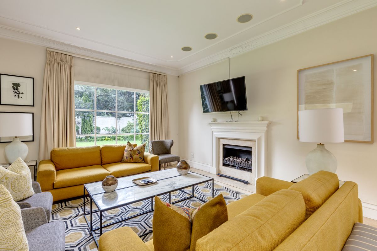 A bright and inviting living room featuring a classic fireplace, decorative crown molding, recessed ceiling speakers, and large windows overlooking the garden.