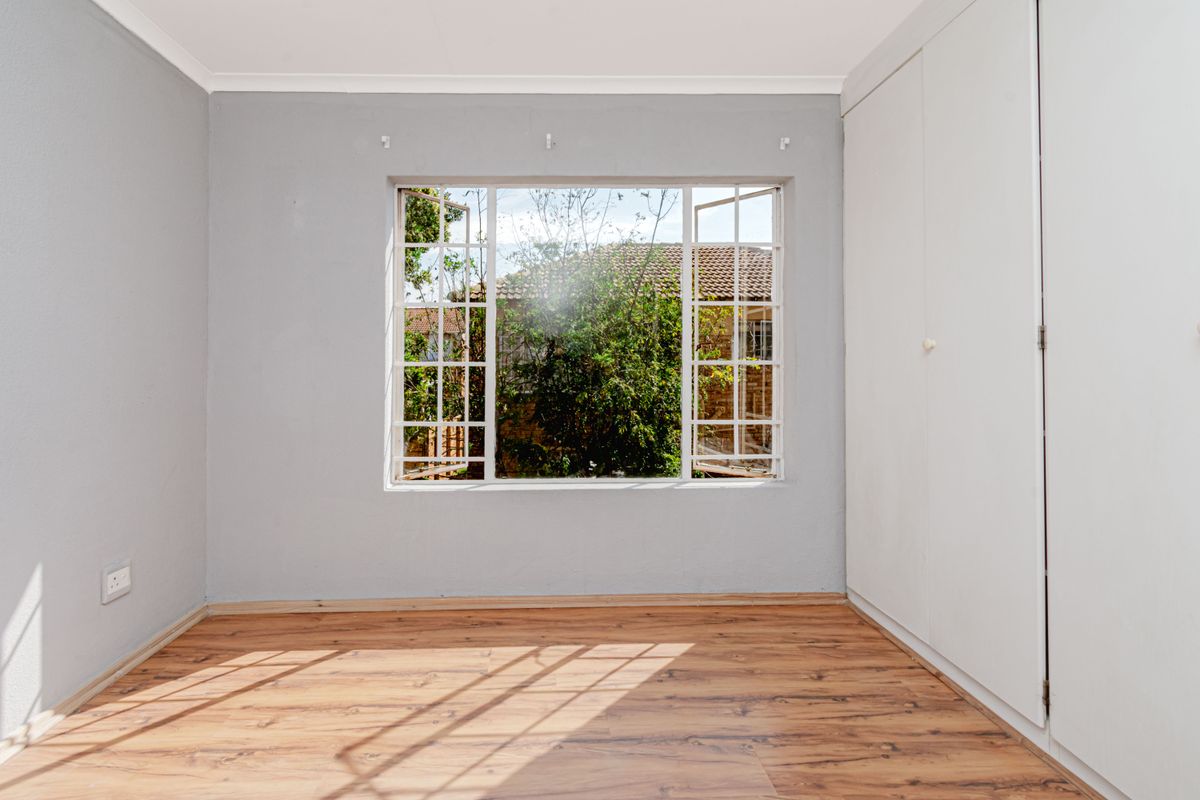 A bright, empty bedroom featuring wood-look laminate flooring, built-in cupboards, and a large window overlooking the garden.