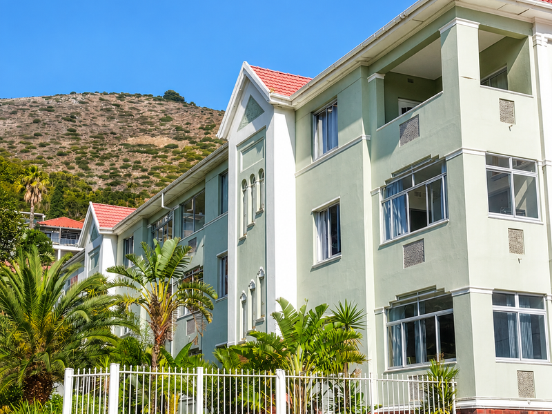 Exterior view of a multi-story apartment building featuring a light green facade, tiled roof, and secure perimeter fencing with landscaped surroundings.