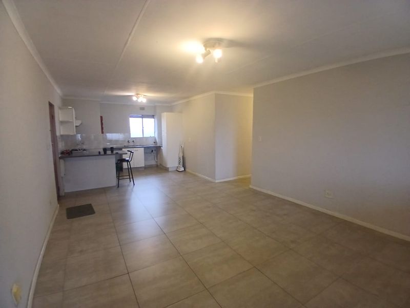 An open-plan living and kitchen area featuring neutral tiled flooring, built-in kitchen cabinetry, and a breakfast counter in a clean, vacant space.