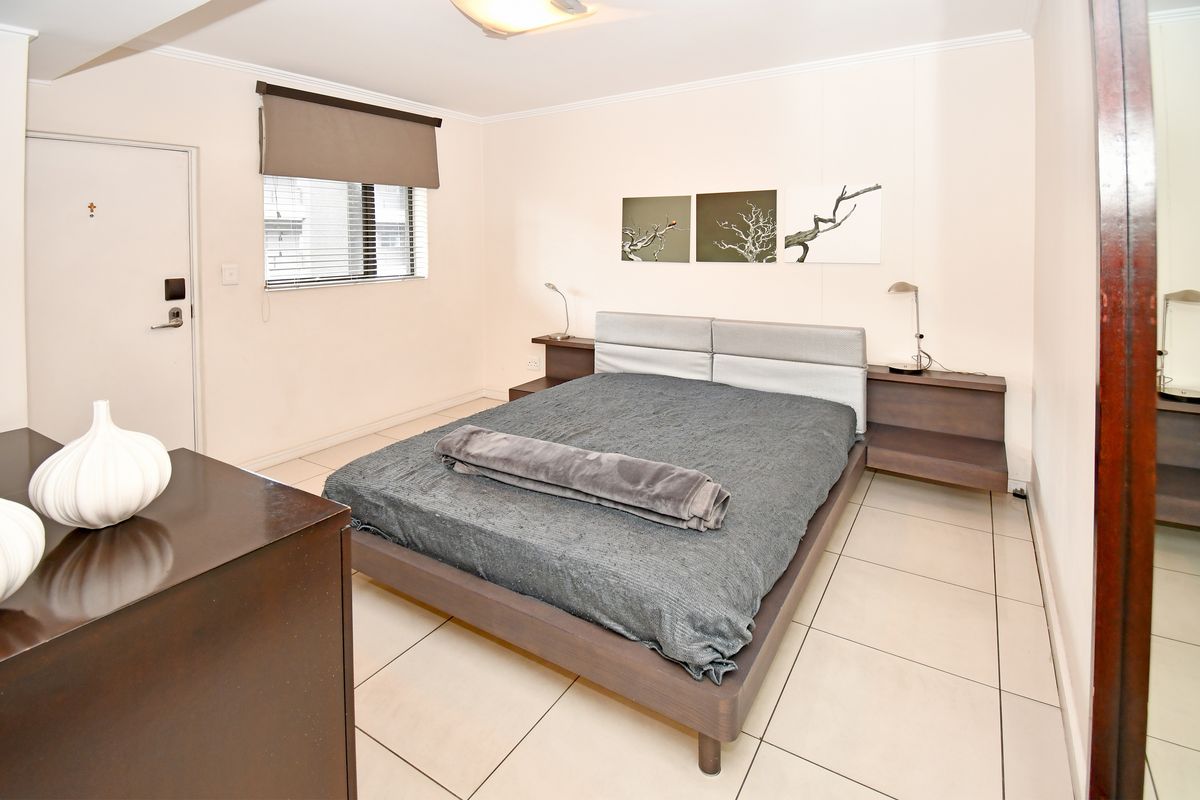 A well-lit bedroom featuring tiled flooring, a built-in headboard with integrated side tables, and a window with a roller blind.