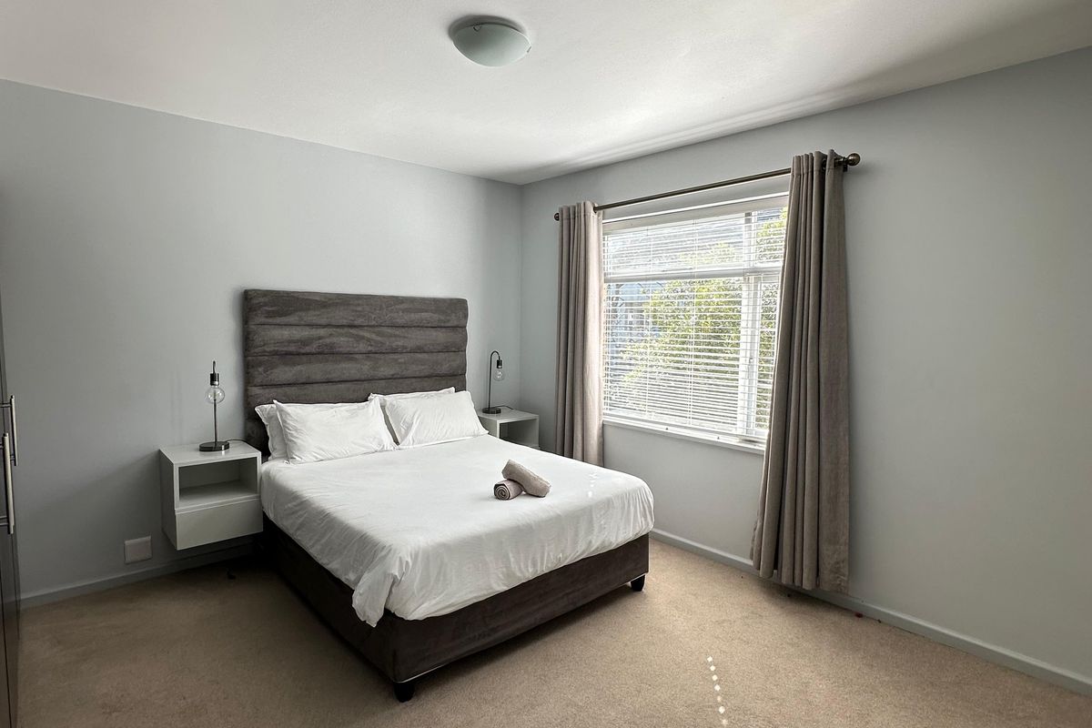 A well-maintained bedroom featuring neutral wall tones, carpeted flooring, and a large window with fitted blinds and curtains.