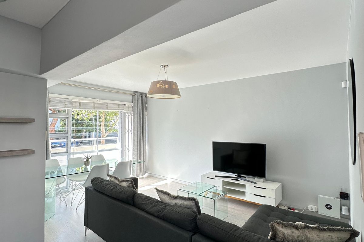 Bright open-plan living and dining area featuring light laminate flooring, neutral walls, and large windows with security bars.