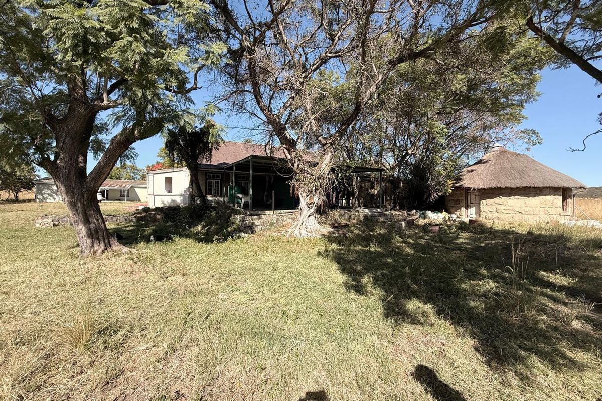Exterior view of a farm property featuring a main dwelling with a covered patio and a traditional thatched-roof outbuilding set in a rural landscape.
