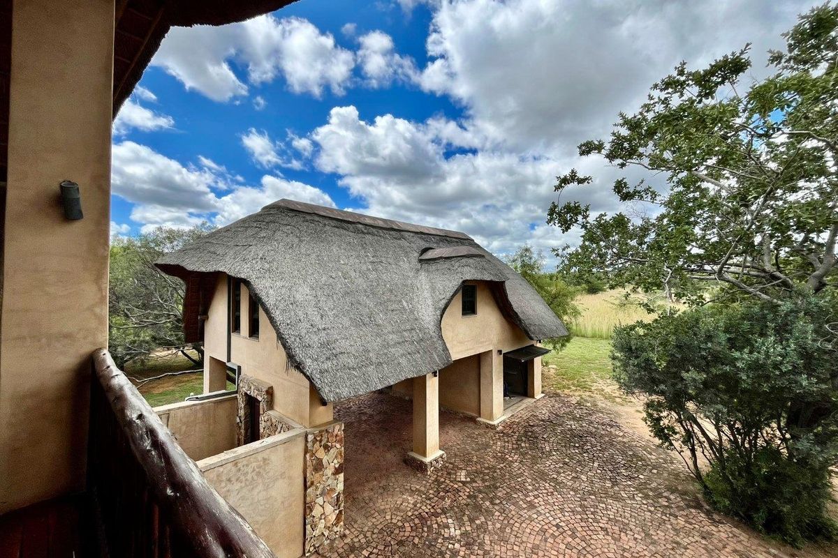Elevated view of a thatched-roof house featuring a paved driveway, stone-clad pillars, and a rustic balcony overlooking the surrounding natural landscape.