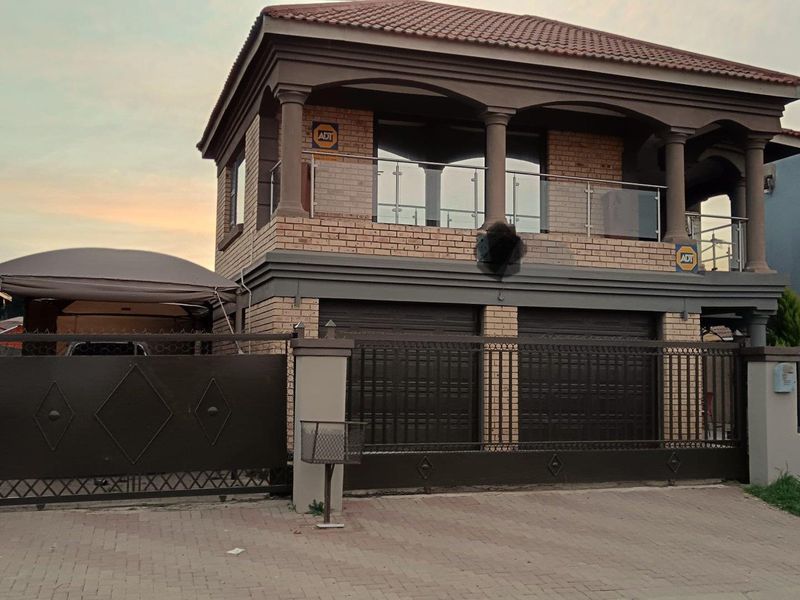 A double-story brick house featuring a double garage, a covered balcony with glass balustrades, and secure metal perimeter fencing.