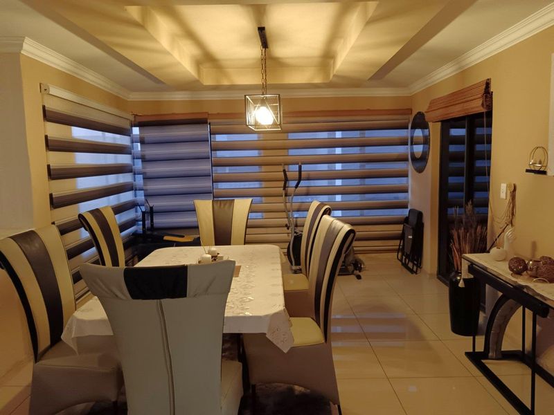 A well-lit dining room featuring a tray ceiling with recessed lighting, modern pendant fixture, tiled flooring, and multiple windows fitted with contemporary roller blinds.
