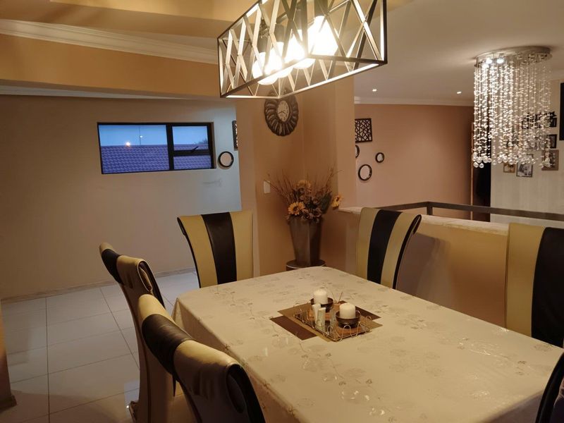 A well-lit dining area featuring tiled floors, a modern geometric pendant light, a crystal chandelier, and an open-plan layout with a half-wall partition.