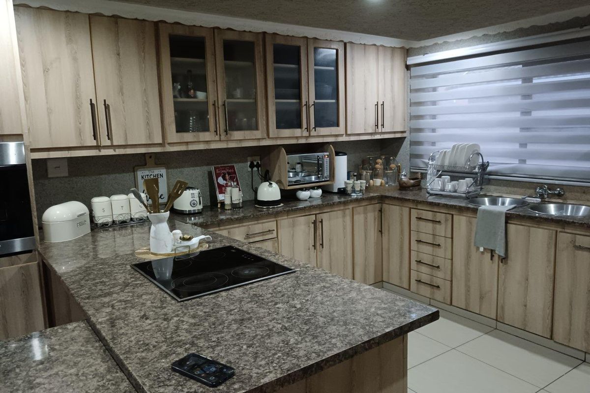 A functional kitchen featuring light wood-tone cabinetry, stone-look countertops, a built-in electric hob, and a double sink with modern roller blinds.