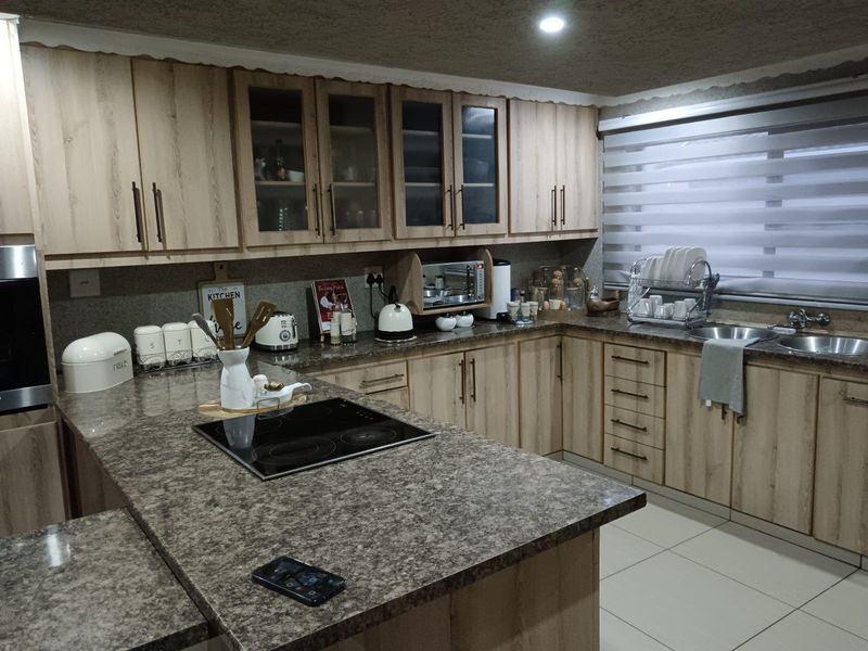 A functional kitchen featuring light wood-tone cabinetry, stone-look countertops, a built-in electric hob, and a double sink with modern roller blinds.