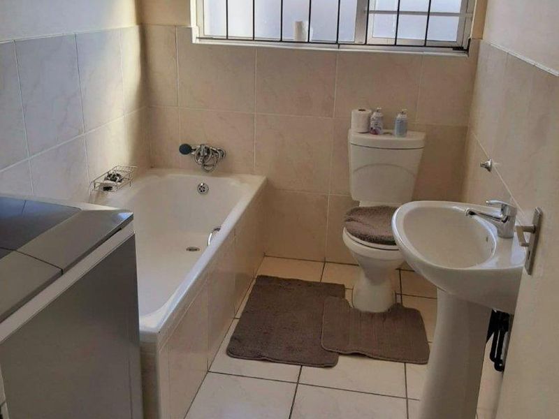 A functional bathroom featuring a bathtub, pedestal basin, and toilet, finished with neutral wall and floor tiles and a security-fitted window.