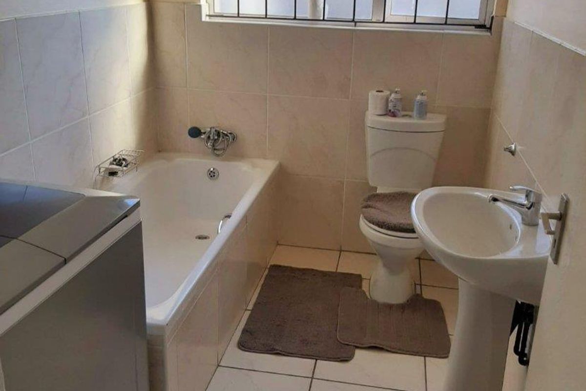 A functional bathroom featuring a bathtub, pedestal basin, and toilet, finished with neutral wall and floor tiles and a security-fitted window.