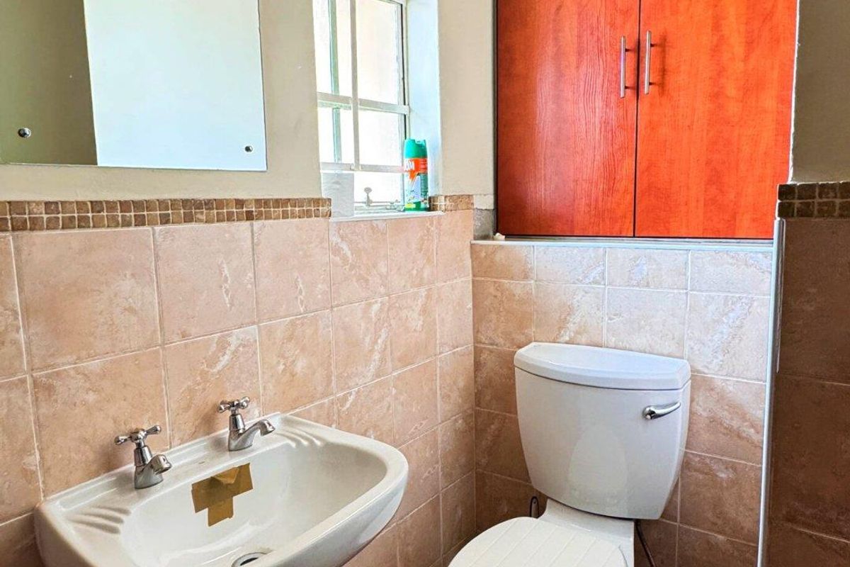 A compact bathroom featuring a pedestal basin, a standard toilet, and built-in wooden cabinetry, finished with neutral wall tiles and terracotta floor tiles.