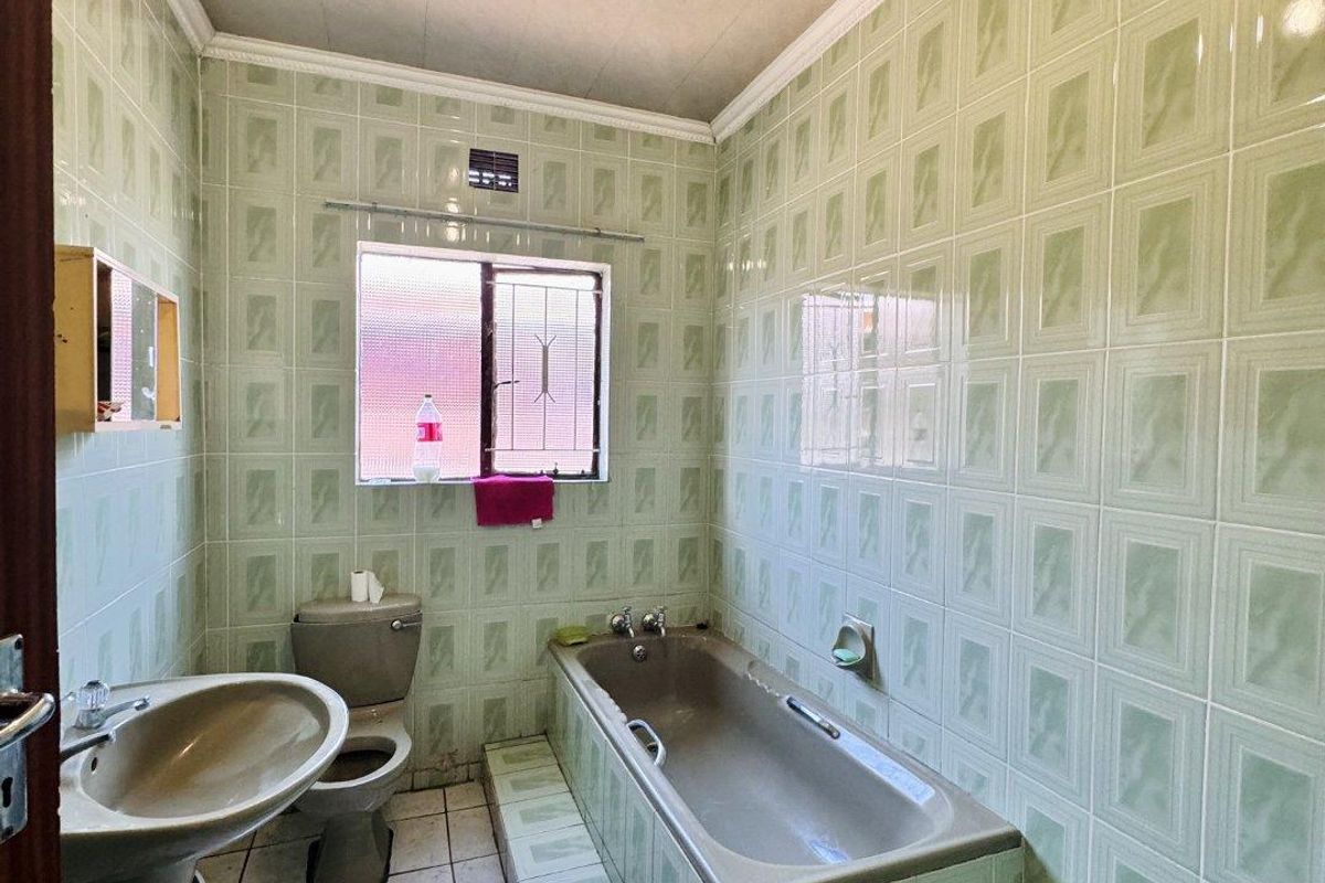 A dated bathroom featuring full-height green wall tiles, a built-in bathtub, pedestal basin, and toilet with tiled flooring.