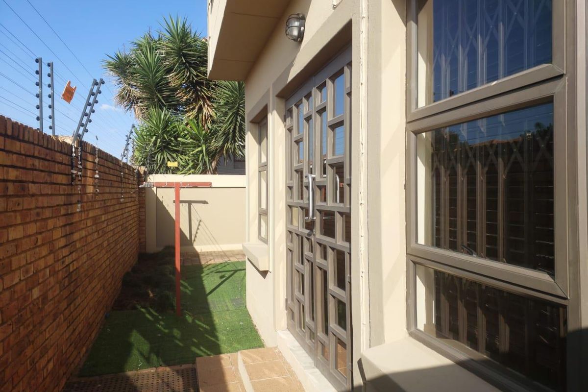 A side exterior view of the property featuring a brick boundary wall, security fencing, and a paved walkway leading to a glass-panelled entrance door.