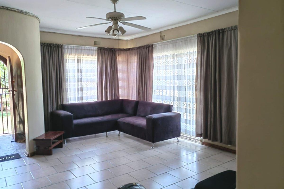 A spacious living room featuring tiled flooring, a ceiling fan, and large windows with curtains, providing access to an adjacent outdoor area.