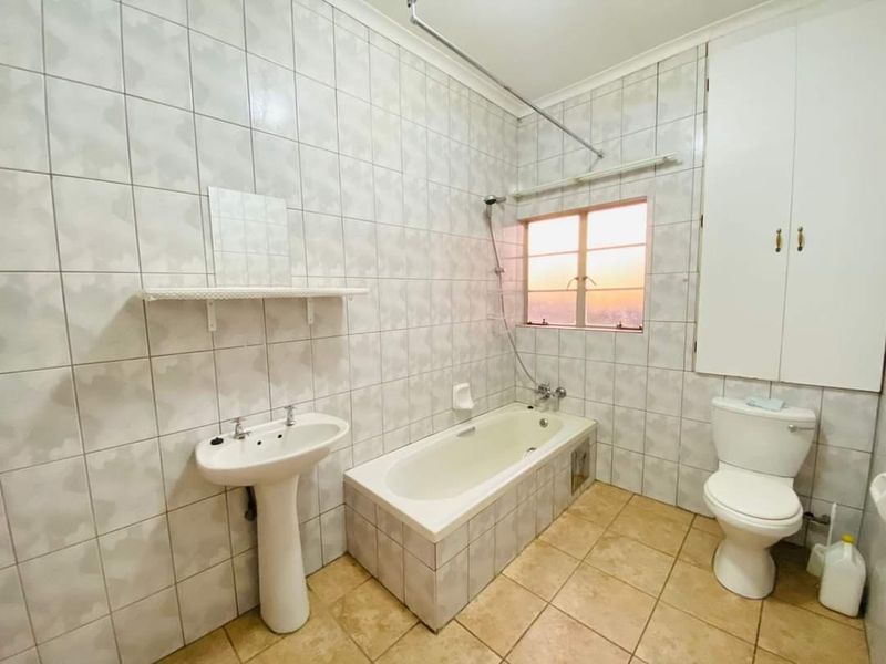 A functional bathroom featuring a pedestal basin, built-in bathtub with shower mixer, toilet, and integrated storage cupboard, finished with tiled walls and flooring.