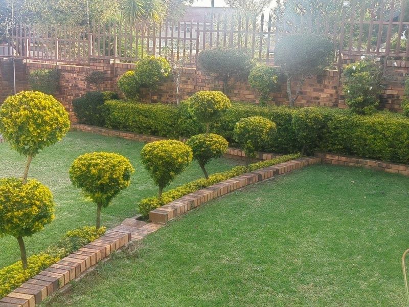 A well-maintained private garden featuring manicured topiary shrubs, a lush lawn, brick edging, and a secure perimeter wall with metal fencing.