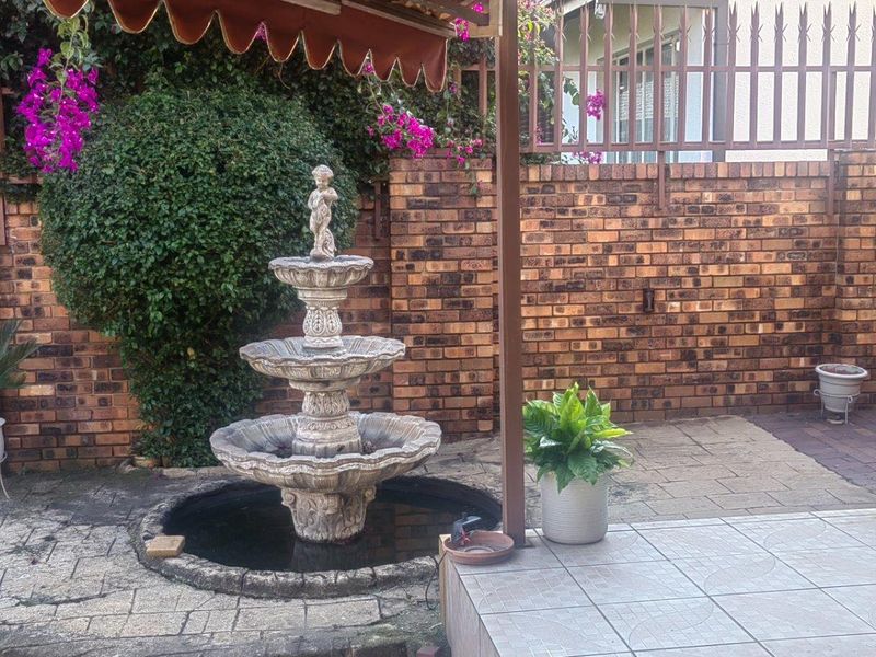 A private outdoor patio area featuring a central stone water fountain, brick boundary walls with security fencing, and a retractable awning for shade.