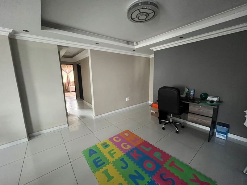 A spacious living area featuring neutral wall tones, tiled flooring, and a recessed ceiling with integrated downlights, leading into an adjacent hallway.