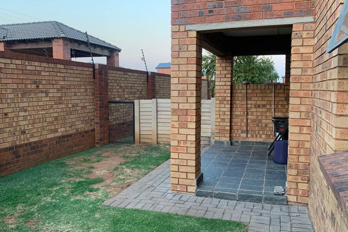 A rear exterior view featuring a covered brick patio with tiled flooring, adjacent to a small garden area enclosed by brick boundary walls.