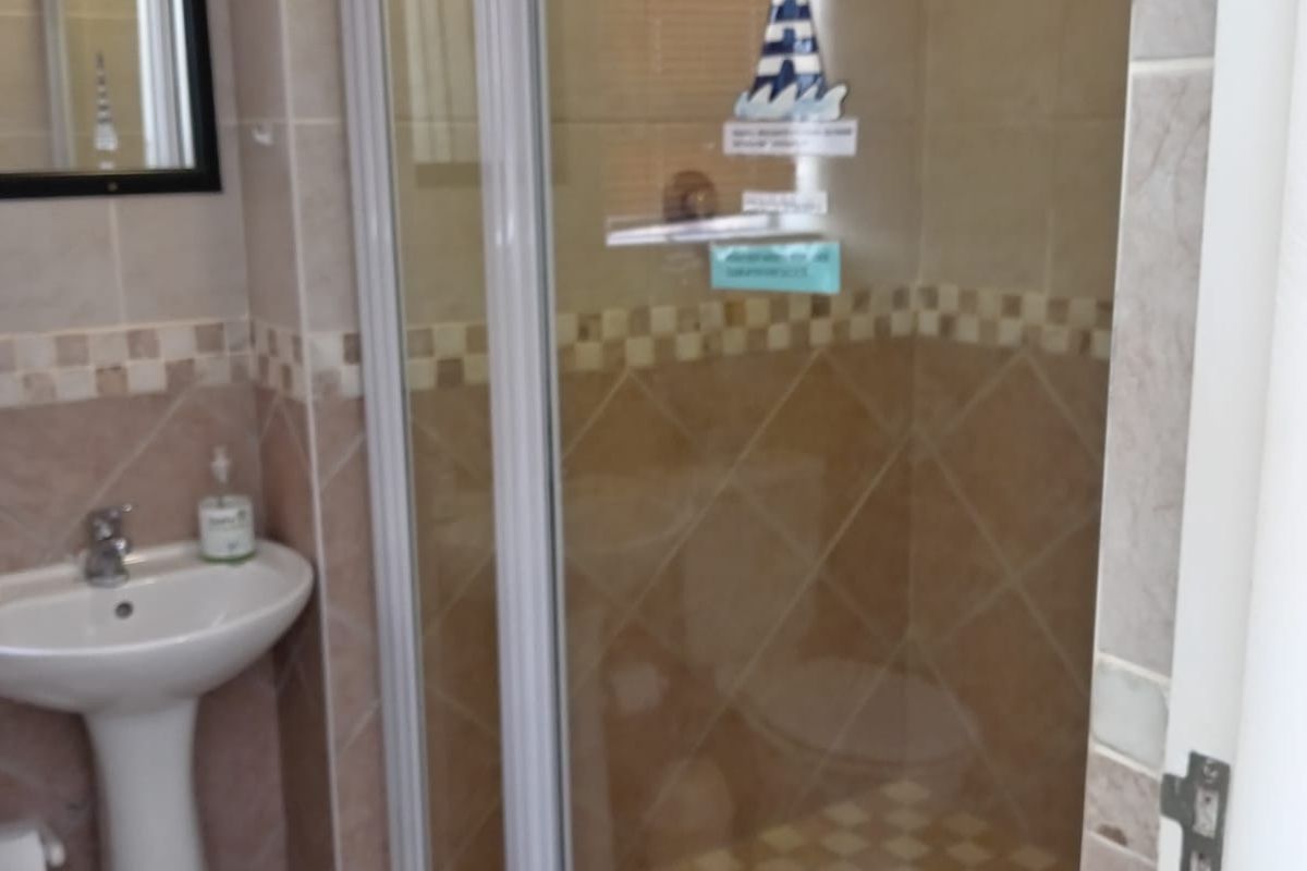 A functional bathroom featuring a pedestal basin, a glass-enclosed shower with tiled walls, and neutral ceramic floor tiling.