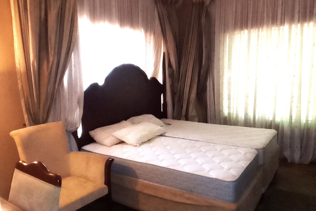 A bedroom featuring large windows with layered curtains, providing natural light. The space is furnished with a bed and an armchair.