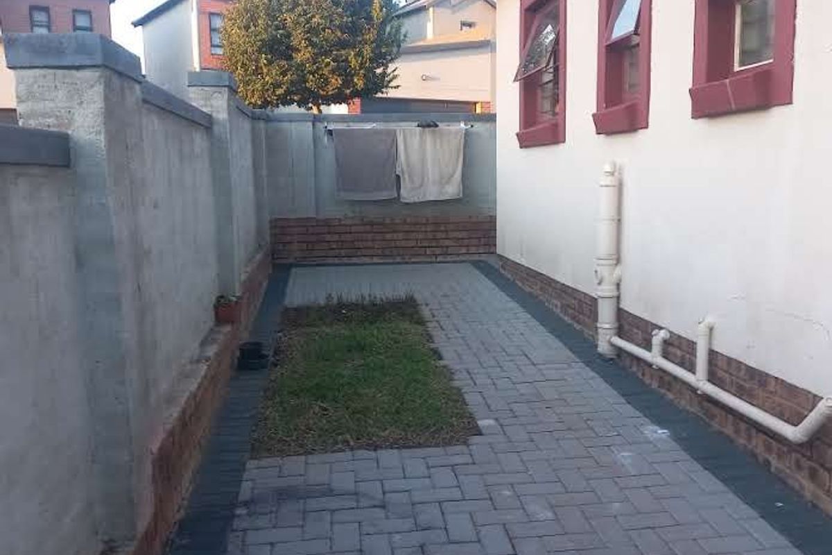 A side exterior view of the property featuring a paved walkway, boundary wall, and exposed exterior plumbing along the building facade.