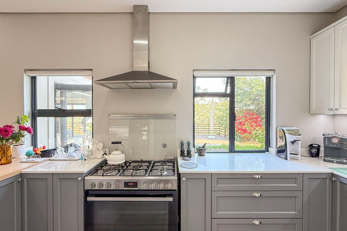 Modern kitchen featuring grey cabinetry, white stone countertops, a stainless steel gas stove with extractor fan, and large windows overlooking the garden.
