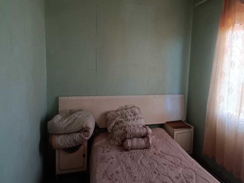 A compact bedroom featuring a built-in headboard and a window with natural light, requiring cosmetic updates to modernize the space.