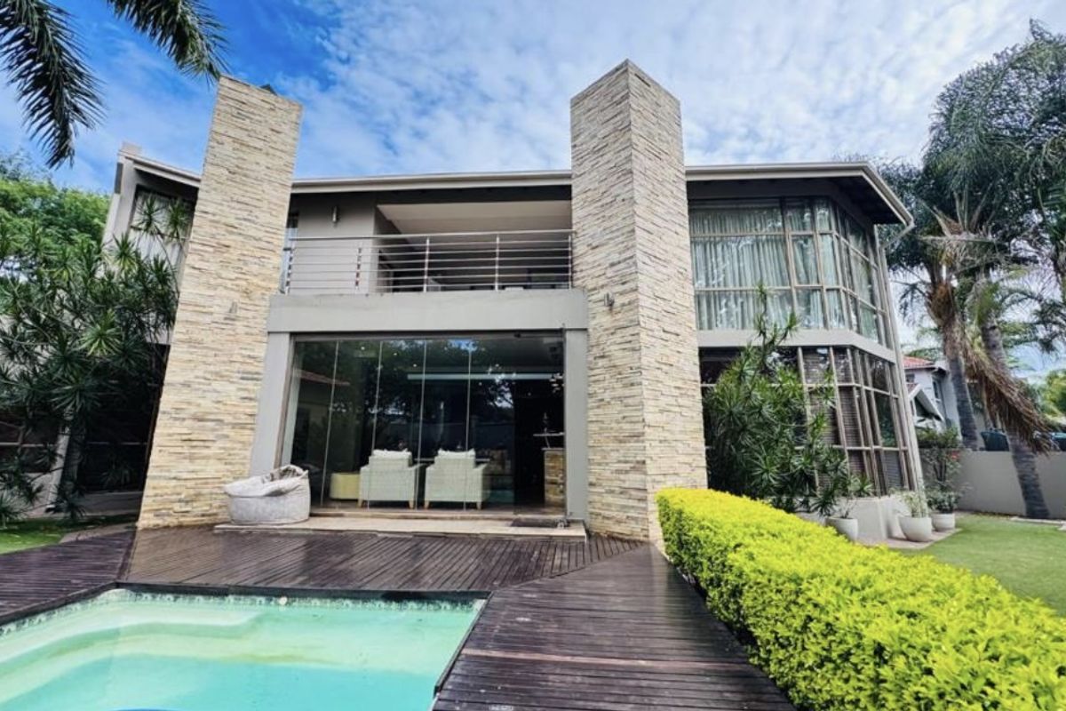 Rear view of a modern double-story residence featuring stone-clad pillars, a glass-enclosed patio, a wooden deck, and a swimming pool.