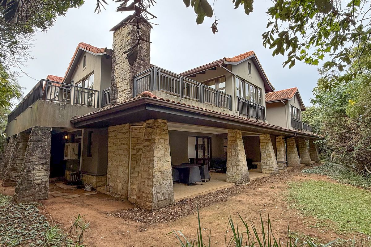 Rear view of a multi-level townhouse featuring stone-clad pillars, a covered patio, an upper-level balcony, and a distinctive stone chimney.