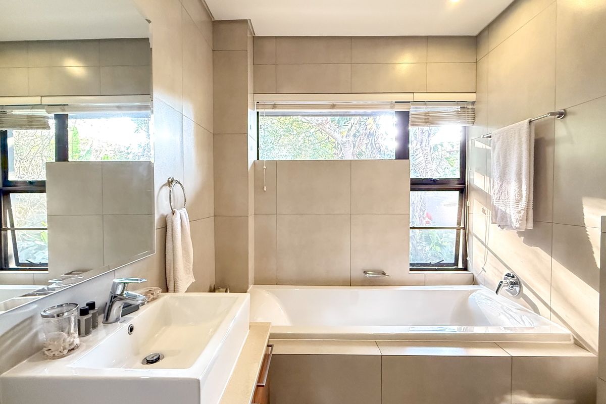 A modern, well-maintained bathroom featuring neutral floor-to-ceiling tiling, a built-in bathtub, and a sleek vanity basin with a large wall-mounted mirror.
