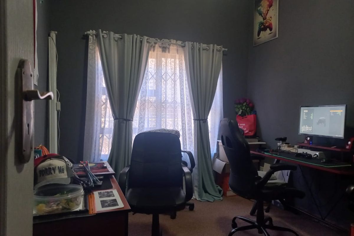 A bedroom currently utilized as a home office, featuring dark-toned walls, carpeted flooring, and a window with curtain treatments.