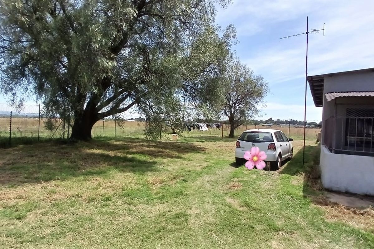 A view of the rear garden area featuring mature trees, a lawn, and a side view of the house structure with security fencing.