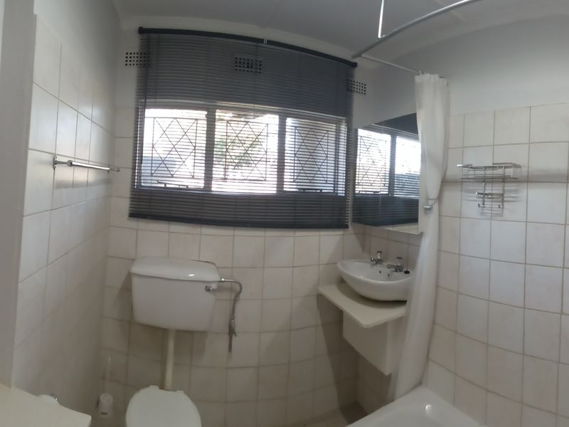 A functional bathroom featuring a bathtub, toilet, and wall-mounted basin with tiled walls and security bars on the window.