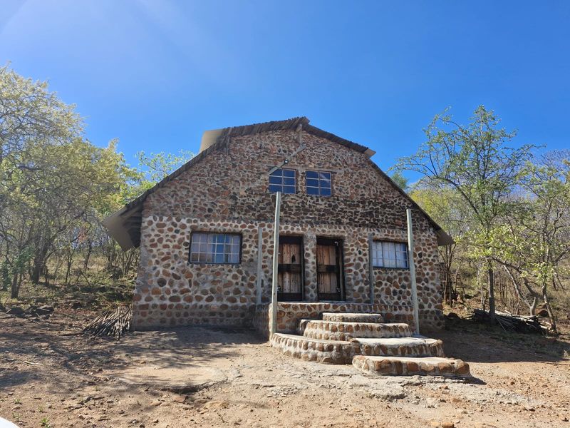 A rustic stone-clad farm cottage featuring a pitched roof, timber entrance doors, and stone steps set within a natural bushveld landscape.