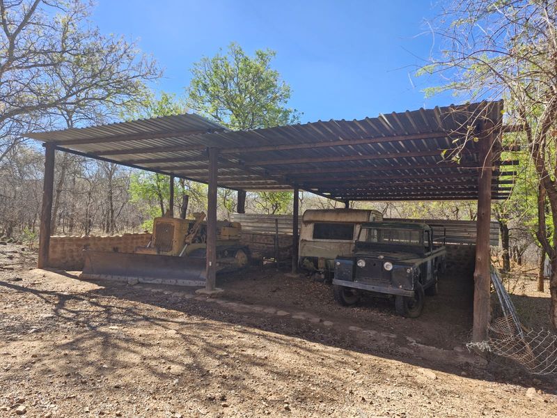 An open-sided farm utility shelter featuring a corrugated metal roof supported by timber posts and a stone retaining wall.