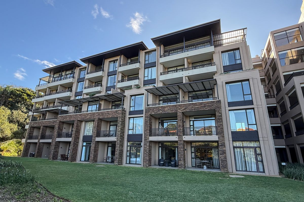 A modern multi-story apartment complex featuring stone-clad pillars, private balconies with metal railings, and large glass windows overlooking a well-maintained lawn.