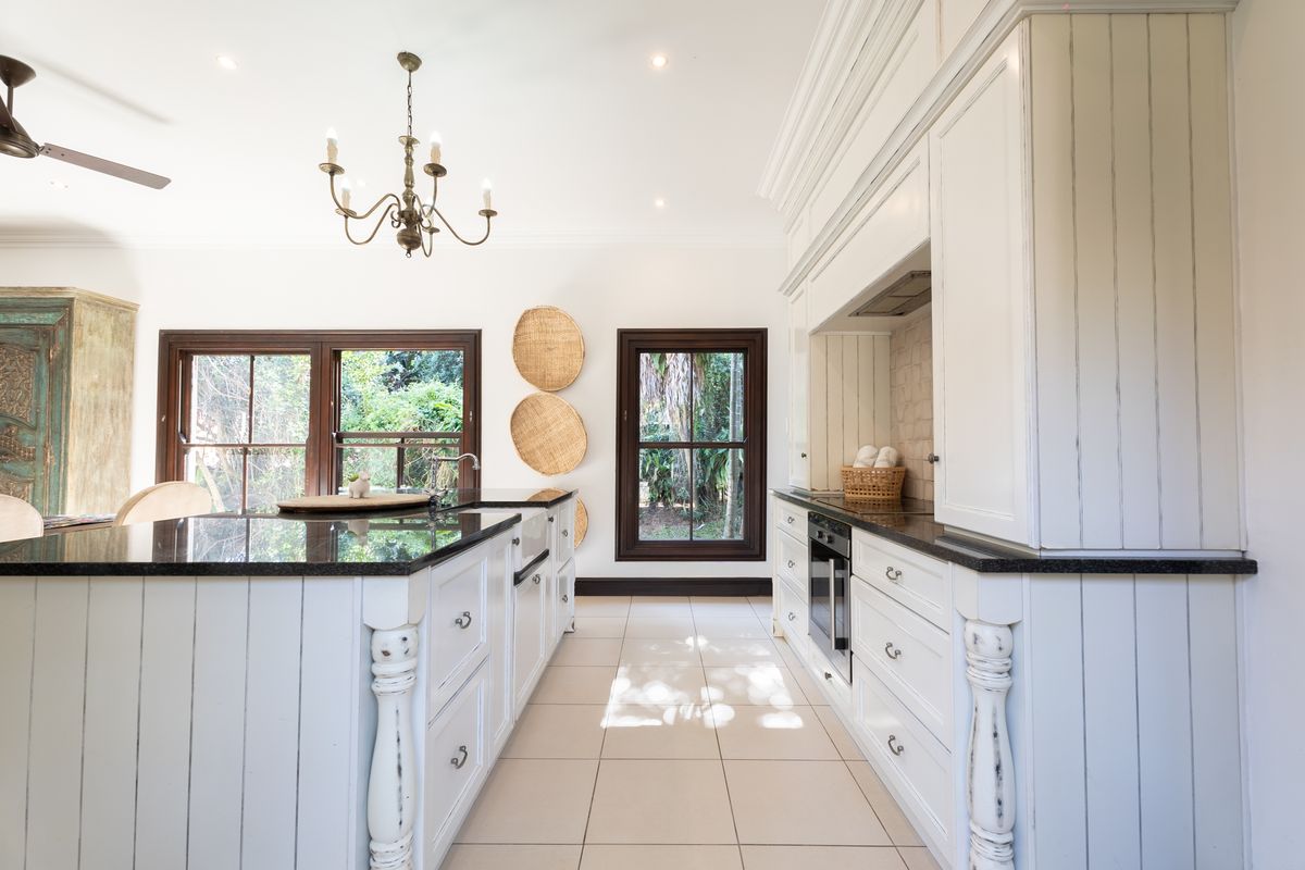 A well-appointed kitchen featuring a large island with granite countertops, farmhouse sink, built-in oven, and classic cabinetry with timber-framed windows overlooking the garden.