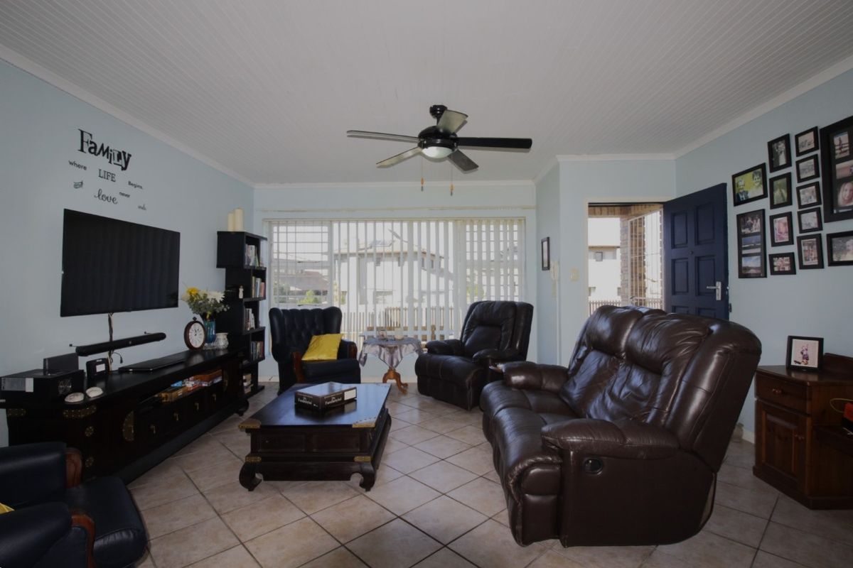 A comfortable living room featuring tiled flooring, a ceiling fan, and large windows with vertical blinds, providing access to the exterior.