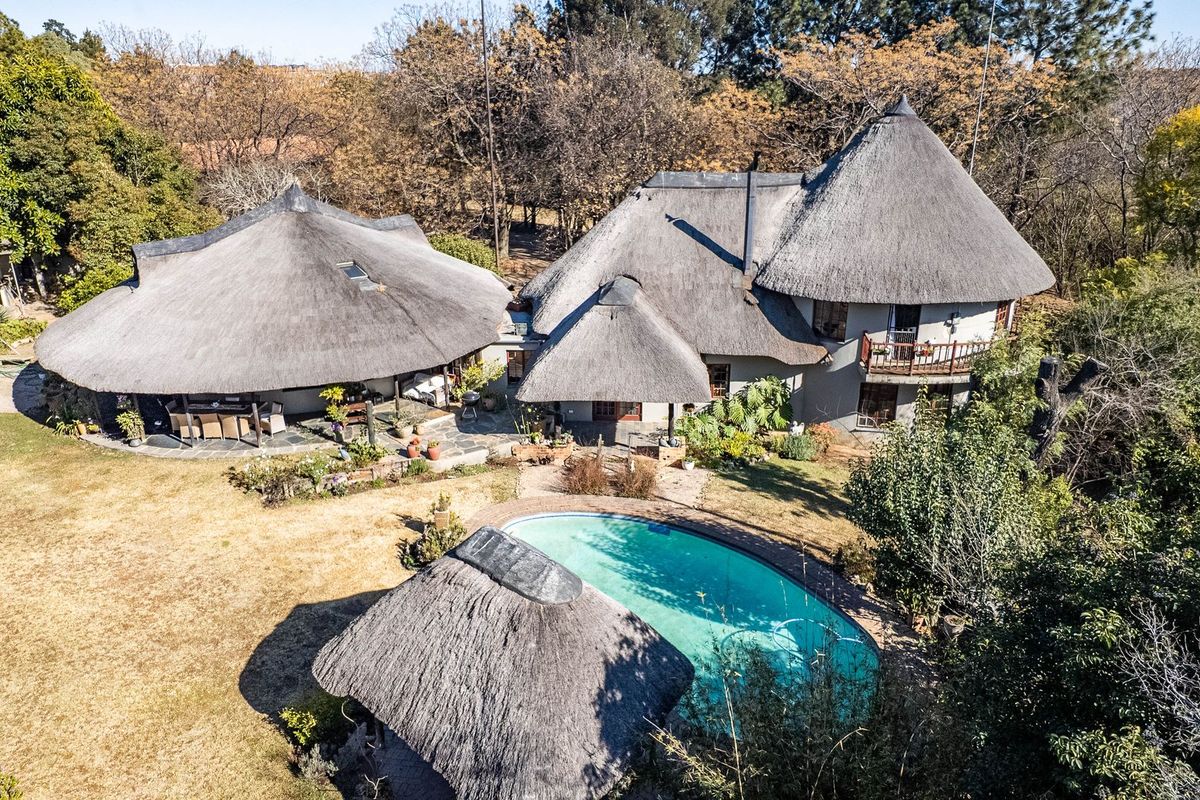 Aerial view of a property featuring multiple thatched roof structures, a swimming pool, stone patio areas, and established garden surroundings.