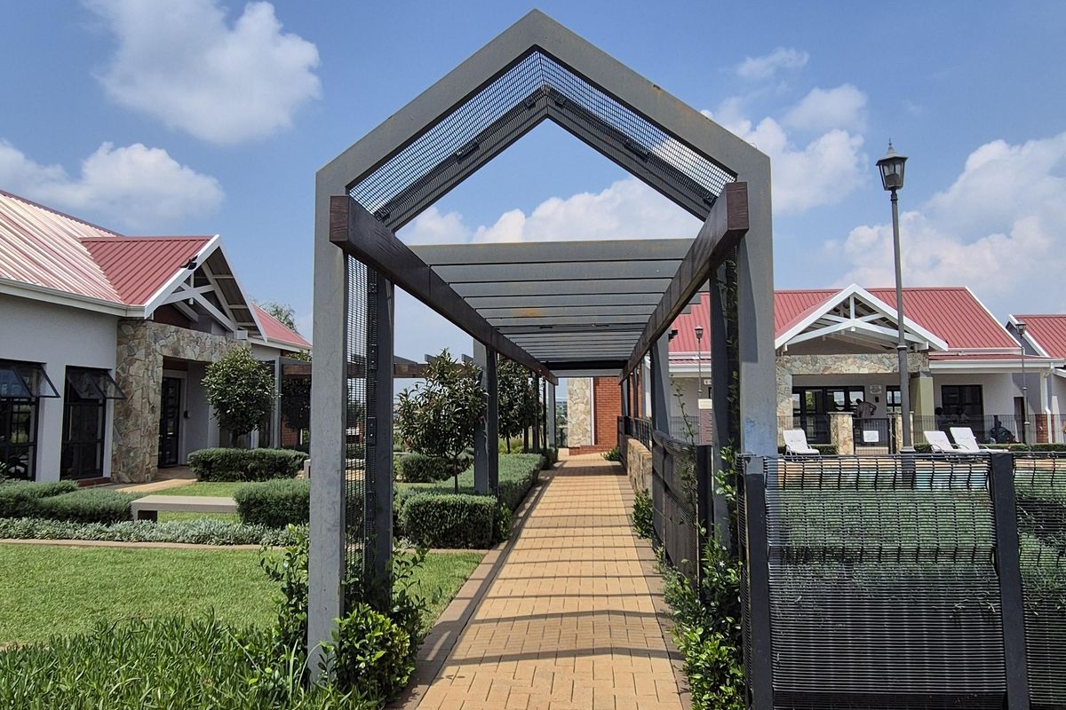 A well-maintained paved walkway featuring a modern metal pergola structure, leading through landscaped garden areas within a residential townhouse complex.